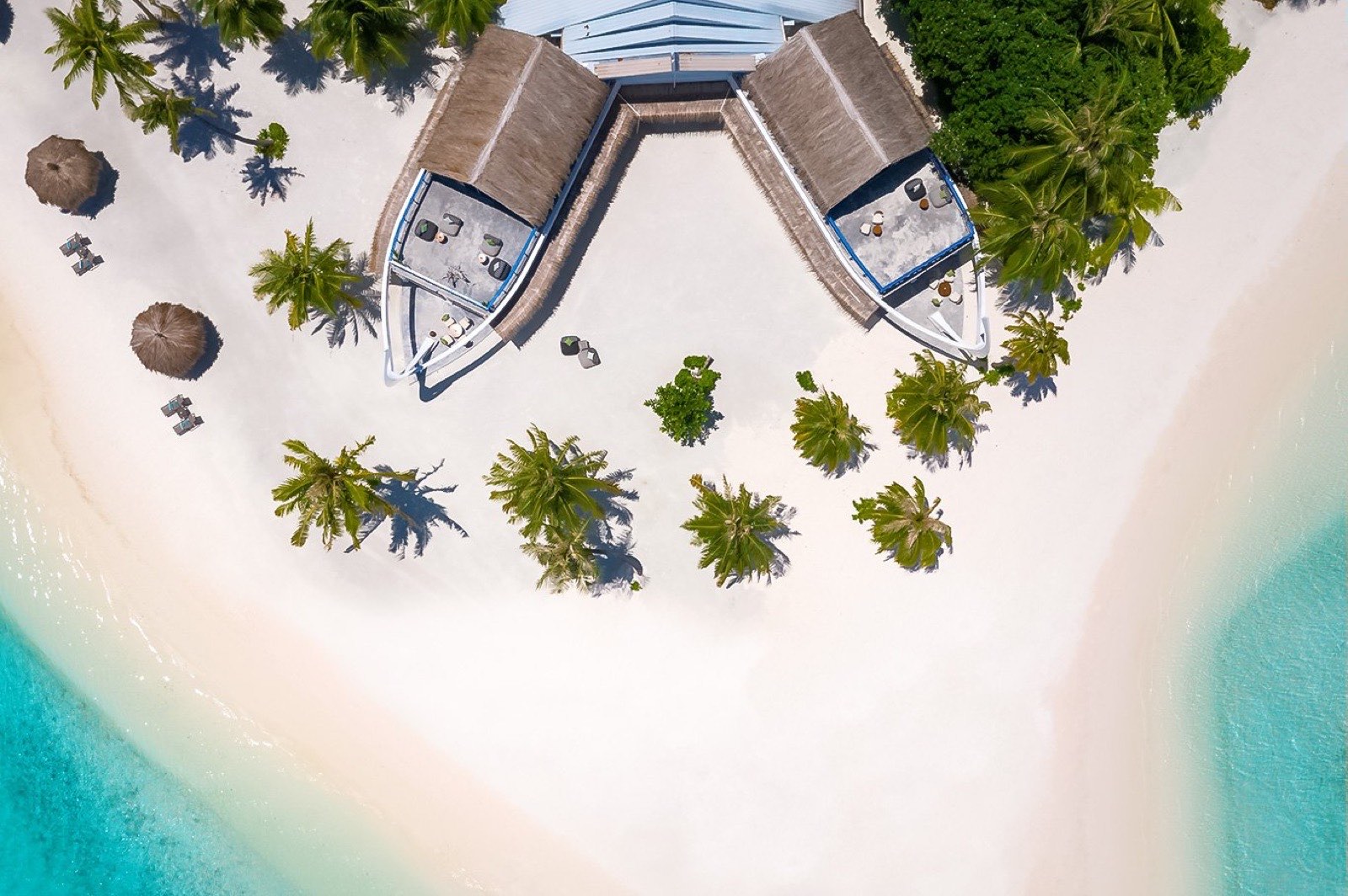 Aerial view straight down over Rahaa Resort: two thatched beach pavilions on wide white sand, palms throwing shadows, turquoise lagoon on both sides.