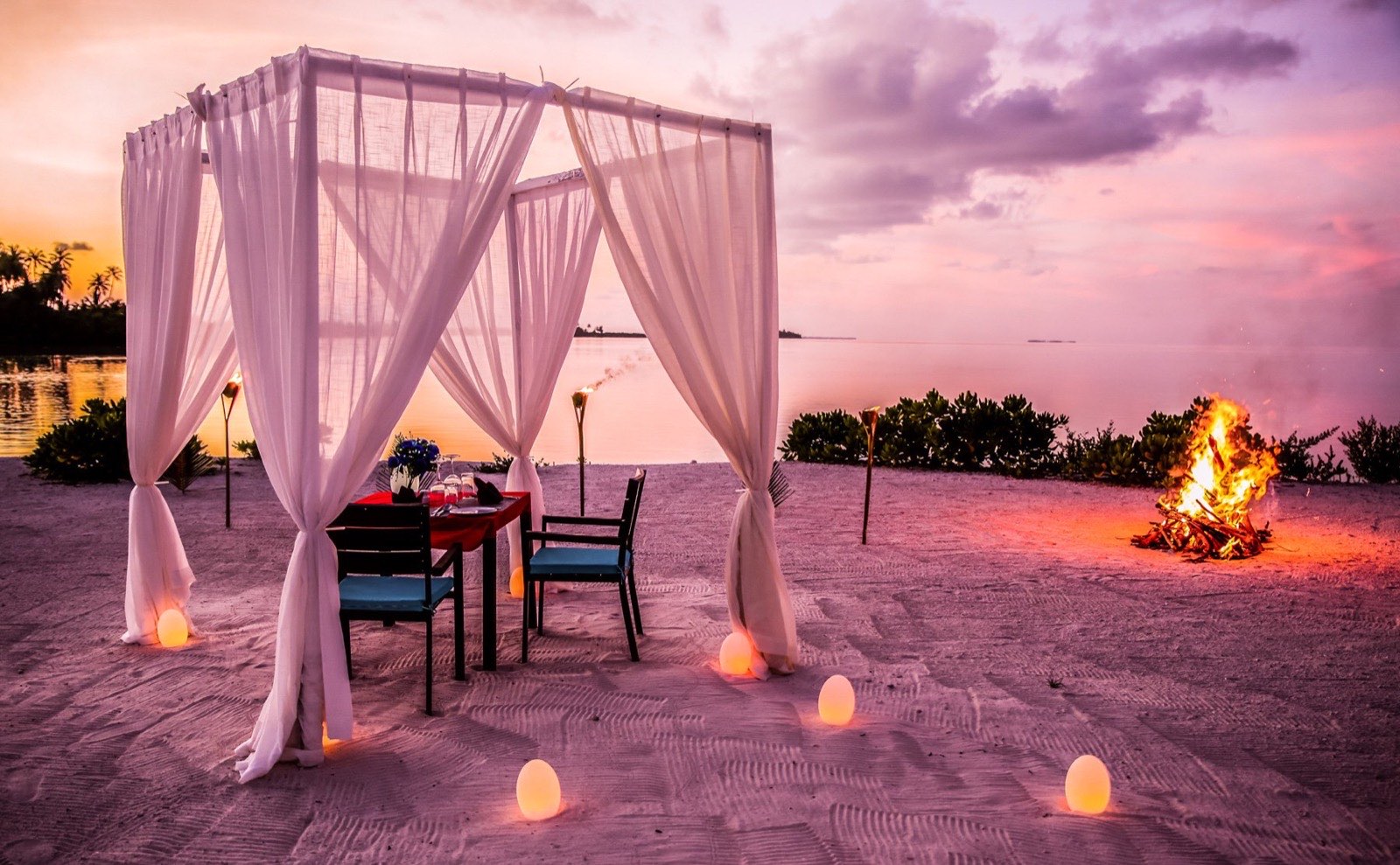 A table set for dinner outdoors on the sand, lit for the evening.